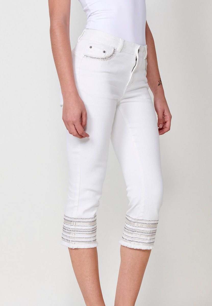 Pantalón jeans capri elástico push up tiro medio y cinco bolsillos con cierre con cremallera y botón metálico y bajo desilachado con bordado cenefa combinado con brillos para mujer de color blanco