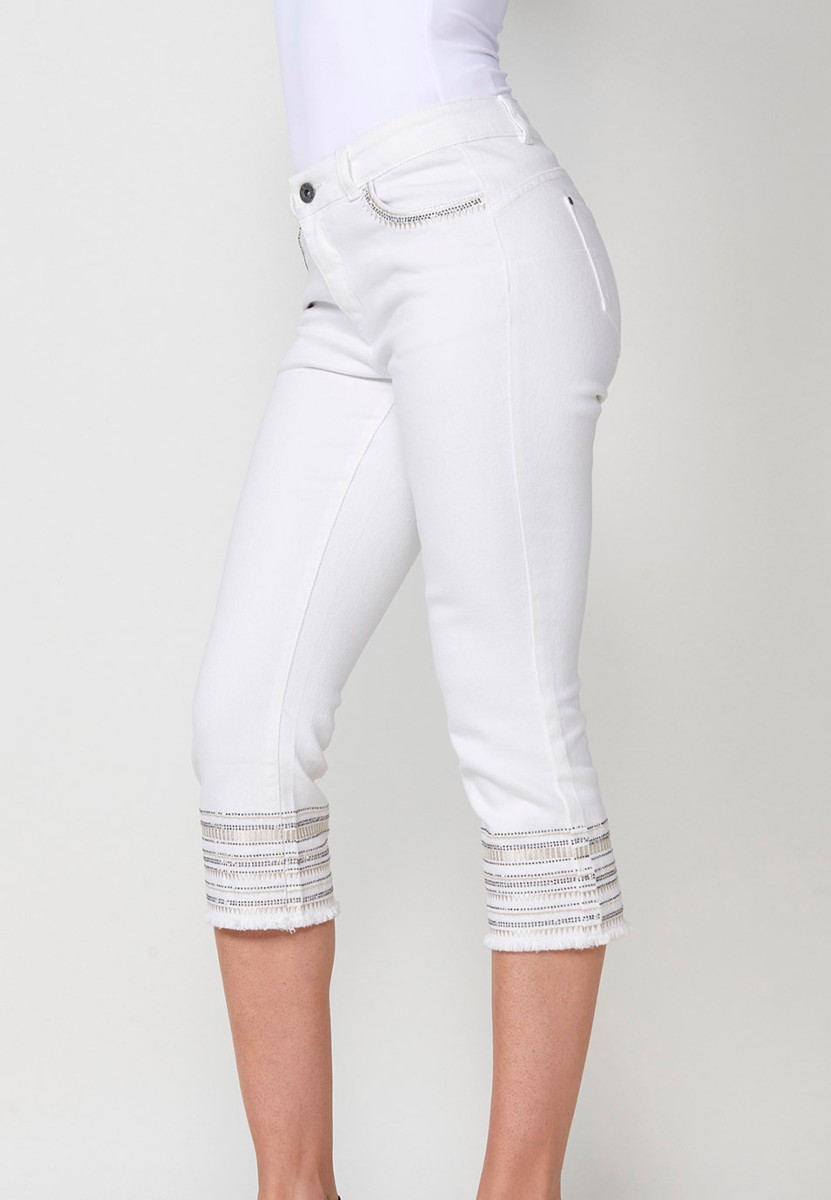 Pantalón jeans capri elástico push up tiro medio y cinco bolsillos con cierre con cremallera y botón metálico y bajo desilachado con bordado cenefa combinado con brillos para mujer de color blanco