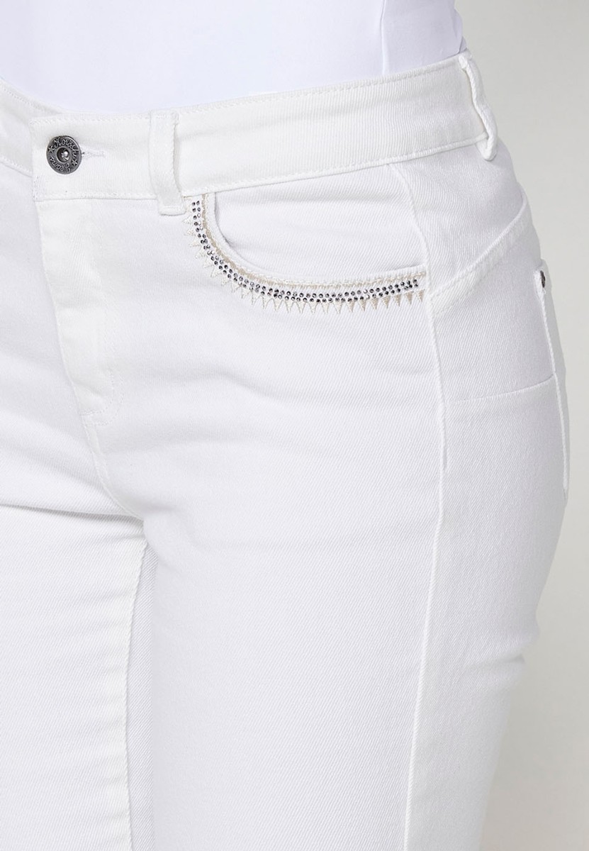 Pantalón jeans capri elástico push up tiro medio y cinco bolsillos con cierre con cremallera y botón metálico y bajo desilachado con bordado cenefa combinado con brillos para mujer de color blanco