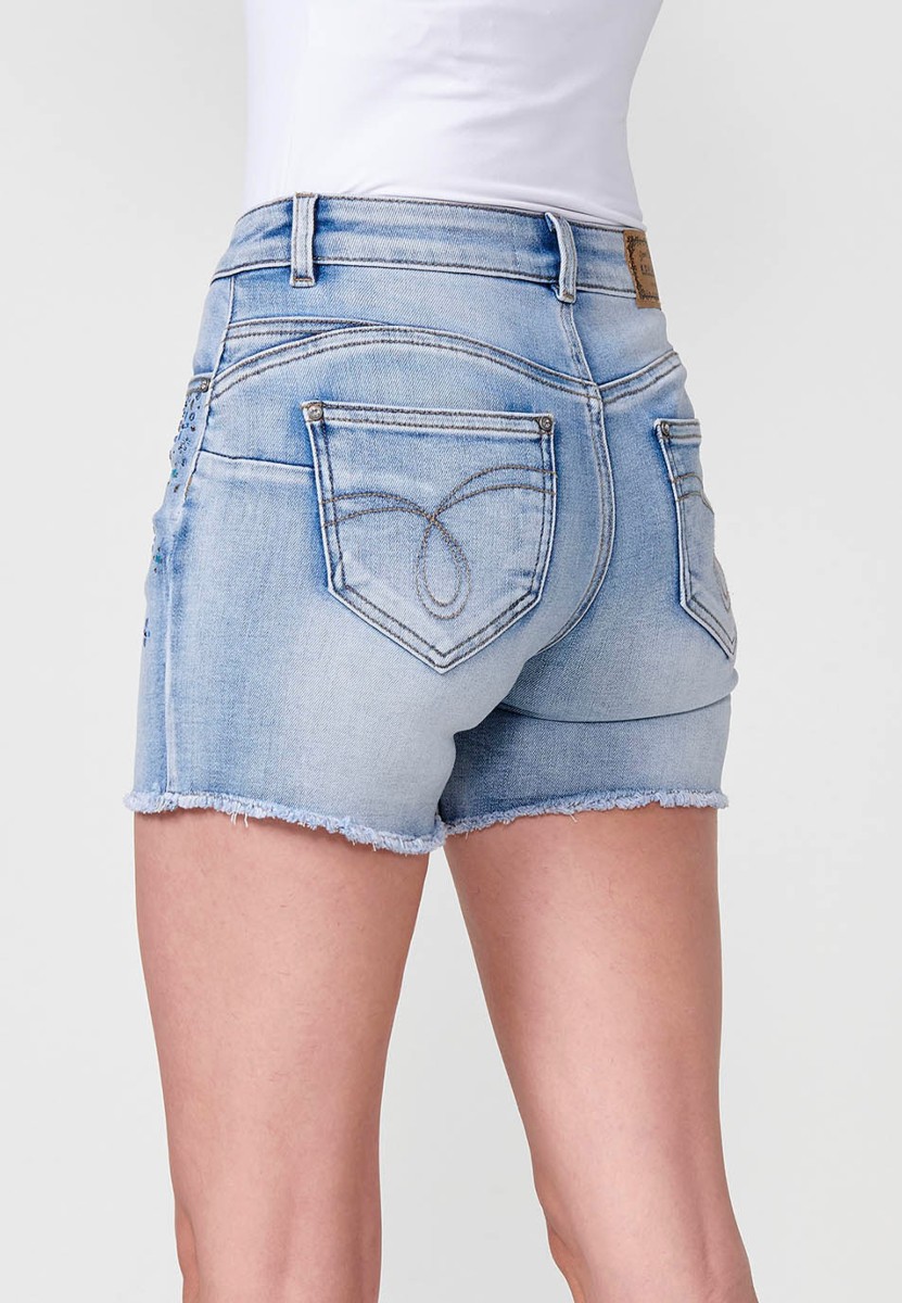 Jeans short elástico push up de tiro medio y cinco bolsillos con cierre y cremallera y botón metálico con detalle de brillos en bolsillos y bajo deshilachado para mujer de color azul claro