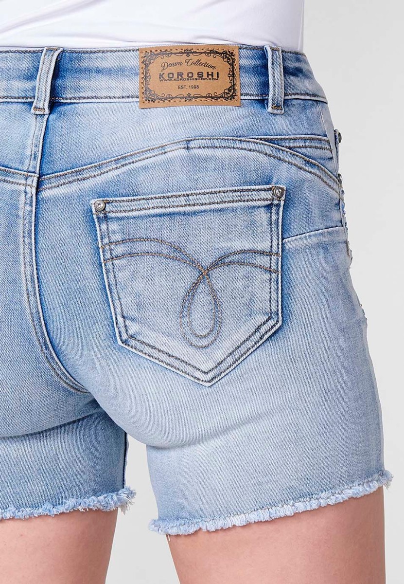 Jeans short elástico push up de tiro medio y cinco bolsillos con cierre y cremallera y botón metálico con detalle de brillos en bolsillos y bajo deshilachado para mujer de color azul claro