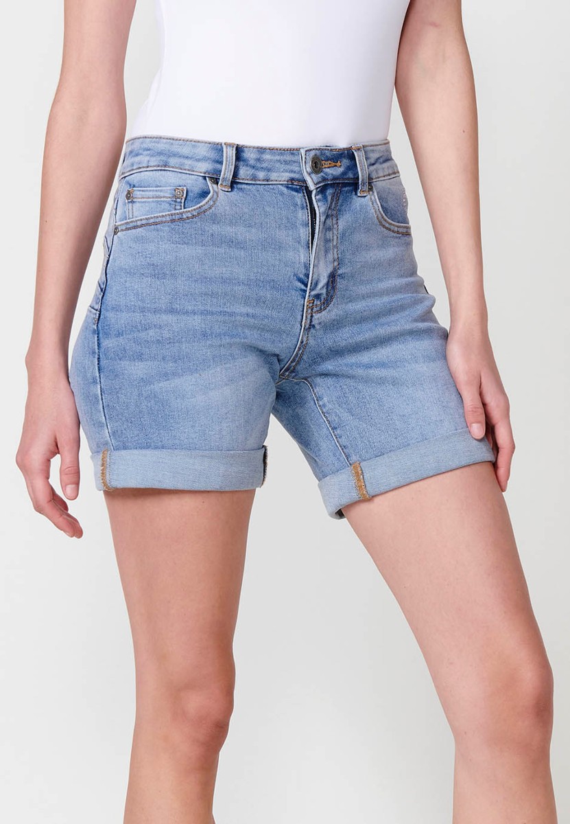 Jeans short elástico de tiro medio y cinco bolsillos con cierre con cremallera y botón metálico con bajo doble vuelta y logo con brillos para mujer de color azul claro