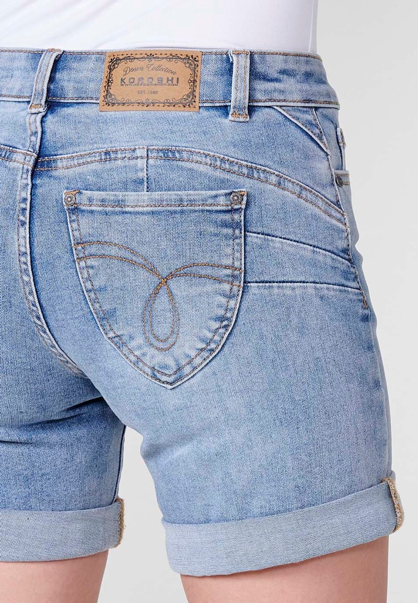 Jeans short elástico de tiro medio y cinco bolsillos con cierre con cremallera y botón metálico con bajo doble vuelta y logo con brillos para mujer de color azul claro