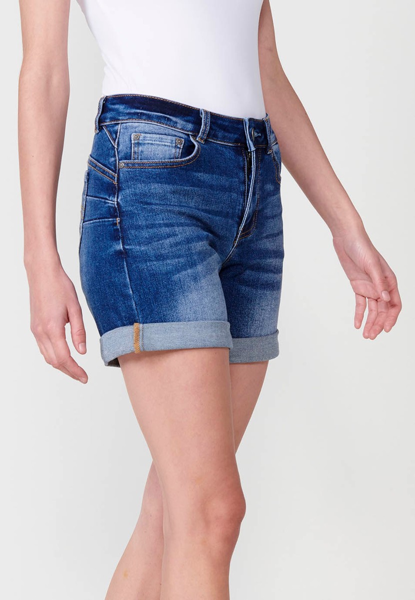 Jeans short elástico de tiro medio y cinco bolsillos con cierre con cremallera y botón metálico con bajo doble vuelta y logo con brillos para mujer de color azul oscuro